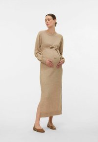 Abito premaman a maglia in un caldo beige, con maniche lunghe, vita aderente con cintura e spacco laterale. Abbinato a semplici scarpe basse marroni.
