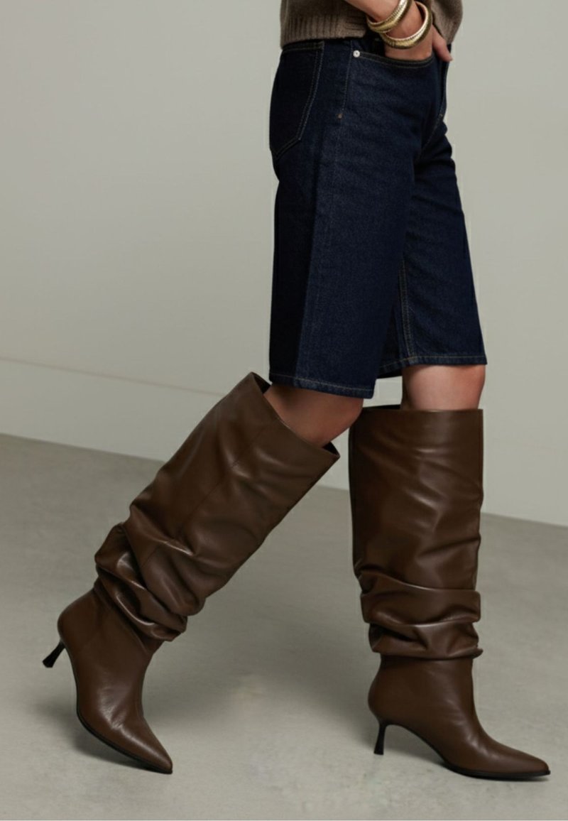 Personne portant un short en jean foncé longueur genou et des bottes à talons en cuir marron décontractées, debout devant un fond uni.