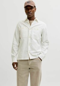 Chemise en coton blanc avec deux poches poitrine, fermeture à boutons et manches longues. Associée à un pantalon beige et une casquette marron.