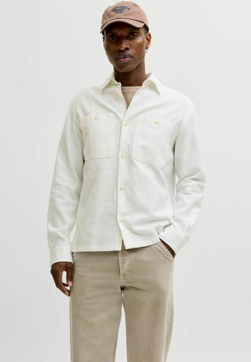 Chemise en coton blanc avec deux poches poitrine, fermeture à boutons et manches longues. Associée à un pantalon beige et une casquette marron.
