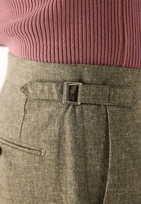 Khaki højttaljede bukser lavet af struktureret stof med en metalspænde og sidelommer, kombineret med en ribstrikket lavendelblå trøje.