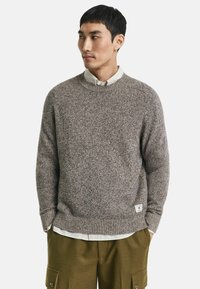 Brauner Strickpullover mit Rundhalsausschnitt, gerippten Bündchen und Saum, strukturiertem Stoff, über einem hellen karierten Hemd getragen, kombiniert mit olivfarbenen Shorts.