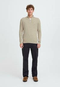 Maglione beige a maglia cable con colletto a mezza zip, abbinato a pantaloni scuri e calzature marroni. Design semplice e strutturato, senza fantasie.