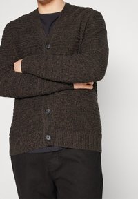 Cardigan lavorato a maglia marrone scuro con motivo testurizzato, scollatura a V e bottoni neri. Indossato sopra una camicia nera, con polsini e orlo a coste.