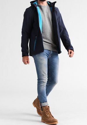 Homme marchant portant une veste bleu foncé avec une doublure bleu vif, un pull gris, un jean bleu clair et des bottes marron à lacets.