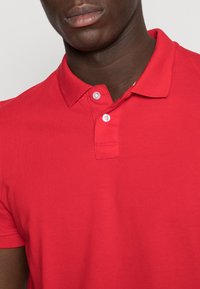 Punainen poolopaita, joka on valmistettu puuvillasta, ja siinä on klassinen kaulus, kaksipainikeinen nappilista ja lyhyet hihat. Rakenne on teksturoitu ja näkyvillä on ompeleet.