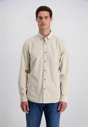 Mann mit kurzen dunklen Haaren, der ein beiges Hemd mit Knöpfen und blaue Jeans trägt, steht vor einem schlichten hellen Hintergrund.