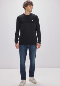Schwarzer Strickpullover mit strukturiertem oberen Bereich und einem Logo-Patch, kombiniert mit blauen Jeans und weißen Sneakers, vor einem schlichten Hintergrund.