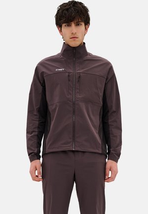 Junger Mann trägt eine dunkelbraune Zip-up-Jacke mit zwei Reißverschlusstaschen auf der Brust und passende Hose vor einfachem hellem Hintergrund.