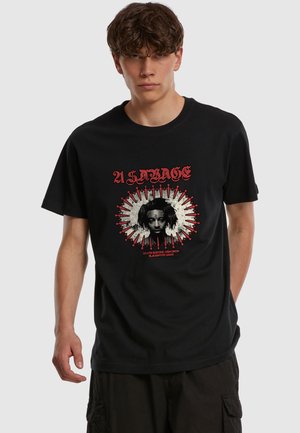Jeune homme portant un t-shirt graphique noir avec un visage central entouré de poignards rouges et un texte gothique « A Savage » sur un fond uni.