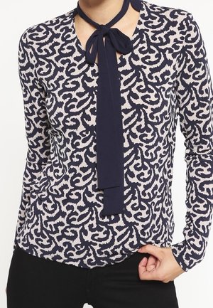 Langærmet marineblå og hvid bluse med abstrakt mønster og bindebånd i marineblåt omkring halsen, båret med sorte bukser.