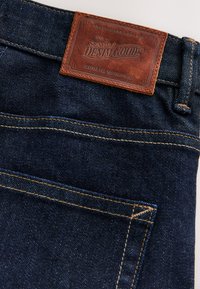 Donkerblauwe denim jeans met zichtbare oranje stiksels en een bruine leren patch met de tekst "Signature Denim Goods" op de tailleband.