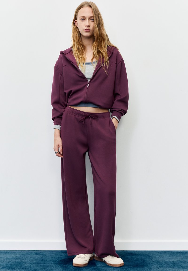 Fioletowa krótka bluza na suwak o gładkiej fakturze, zestawiona z pasującymi szerokimi spodniami dresowymi. Widoczna szara warstwa wewnętrzna pod bluzą. Jasne sneakersy.