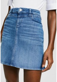 Femme portant une mini-jupe en jean bleu moyen avec des poches devant et une chemise blanche rentrée, debout devant un fond uni.