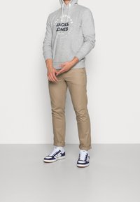 Sudadera gris con cordones blancos y logotipo negro, combinada con pantalones tapered color beige y zapatillas deportivas azul marino con suelas y cordones blancos.