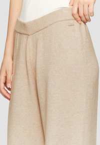 Beige gebreide broek met een geribbelde tailleband, voorzien van zijzakken en een gestructureerde stof. Subtiele kleurvariaties zichtbaar.