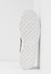 Semelle d'une chaussure blanche Clarks avec des motifs de bande de roulement circulaires et ondulés, inclinée contre un coin de mur blanc, montrant un bord supérieur à imprimé léopard.