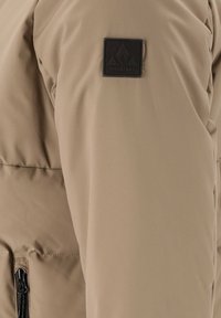 Beige-päällysteinen takki, jonka hihassa on musta Whistler-logo-patch ja musta vetoketjullinen taskudetaili.