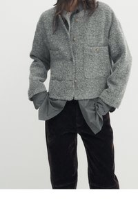 Veste en mélange de laine grise avec une finition texturée, dot�ée de deux poches avant et d'une fermeture à bouton, portée par-dessus une chemise gris clair et un pantalon marron foncé.