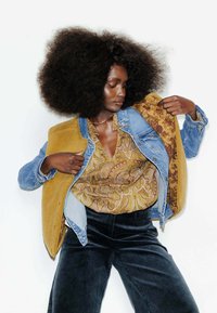 Veste en denim avec doublure moutarde, intérieur floral et blouse dorée texturée. Le mannequin porte un pantalon en velours côtelé noir, mettant en valeur des motifs vibrants.
