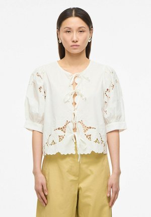 Naine, kes kannab valget pitsist pluusi lipsueesriidega ja puffy varrukatega, kombineeritud kõrge vöökohaga beežide pükstega, seistes valge tausta ees.