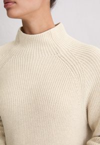 Kremowy sweter dzianinowy z wysokim kołnierzem, z fakturowanym wzorem i rękawami raglanowymi. Materiał wydaje się miękki i przewiewny.