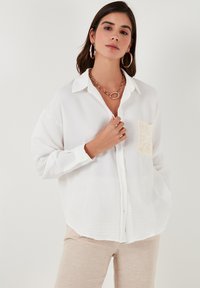 Camisa blanca de botones con una tela texturizada, mangas largas y un gran detalle de bolsillo de ganchillo en el lado izquierdo.