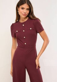Bordó jumpsuit rövid ujjakkal, két felső zsebbel és díszes ezüst gombokkal az elején. Sima anyag és testhezálló kialakítás.