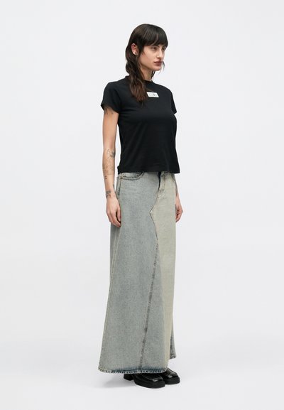 Žena oblečená v čiernom tričku s krátkym rukávom a dlhom, dvojfarebnom denimovom sukni s vysokým pásom, s �čiernymi topánkami, stojaca pred bielym pozadím.