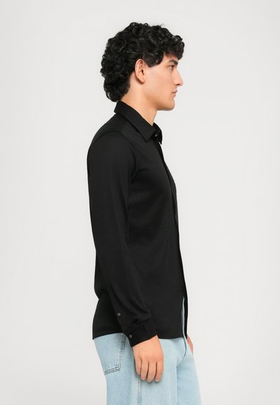 Mann med krøllete hår iført en svart langermet skjorte med knapper og lys blå jeans, stående i profil mot en hvit bakgrunn.