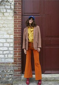 Blazer à carreaux, chemise à motifs jaunes, pantalons orange évasés, bottines rouges et casquette bleue. Matériaux texturés avec des boutons sur les pantalons, pose debout.
