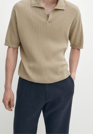 Mann trägt beiges geripptes kurzärmeliges Strick-Poloshirt und dunkelblaue Hose, steht mit einer Hand in der Tasche vor einfarbigem Hintergrund.