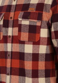Rot und orange kariertes Flanellhemd mit einem Hahnentritt-Muster, zwei Brusttaschen und braunen Knöpfen. Der Stoff hat eine weiche Textur.