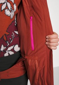 Veste rouge avec une poche zippée rose et une texture lisse. Fermeture à bouton visible en bas, avec un design en tissu superposé.