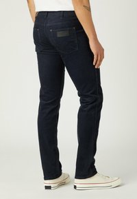 Navy denim jeans with a slim fit, featuring two back pockets and a leather logo patch. Paired with white low-top sneakers.