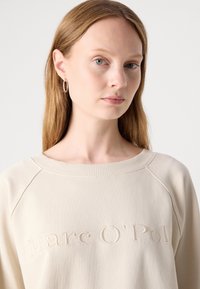 Felpa beige con scollatura a costine e ricamo del testo "Marc O’Pol" sul davanti. Tessuto morbido e vestibilità relaxed con maniche a raglan.
