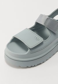 Lyseblå sandaler med en tykk såle, justerbar grå stropp, teksturert fotseng og UGG-logo på siden. Rund design med åpen tå.