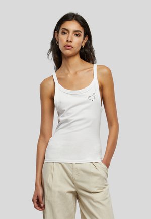 Femme portant un haut blanc sans manches avec un petit motif de visage de chien sur la poitrine et un pantalon beige, debout avec une main dans la poche.
