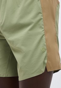 Κοντινό πλάνο σε σορτς ελιάς και καμηλό με μικρό στρογγυλό λογότυπο στο πλάι, φορεμένα από άτομο με σκούρο δέρμα.