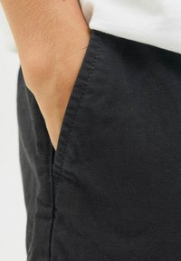 Tissu noir avec une finition texturée ; une main repose dans la poche, révélant un design simple aux lignes épurées et aux détails de couture subtils.