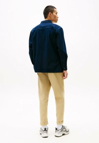 Camicia blu navy con colletto abbottonato e maniche lunghe, abbinata a pantaloni chinos beige e sneakers bianche con accenti neri.