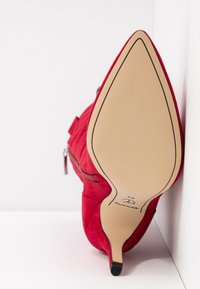 Chaussure à talon aigu en daim rouge avec un bout pointu, dotée d'une fermeture éclair sur le côté et d'une semelle beige avec un logo rond au talon.