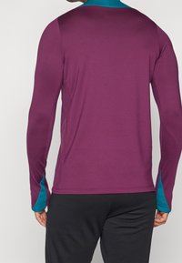 Długi rękaw, sportowa bluzka w głębokim purpurze z turkusowym akcentem na mankietach. Gładki, elastyczny materiał z dopasowanym krojem i bocznymi rozporkami.