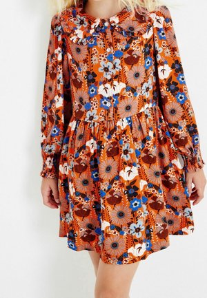 Fille portant une robe orange à manches longues avec des motifs floraux bleus, blancs et bruns et un col à volants, debout devant un fond blanc.