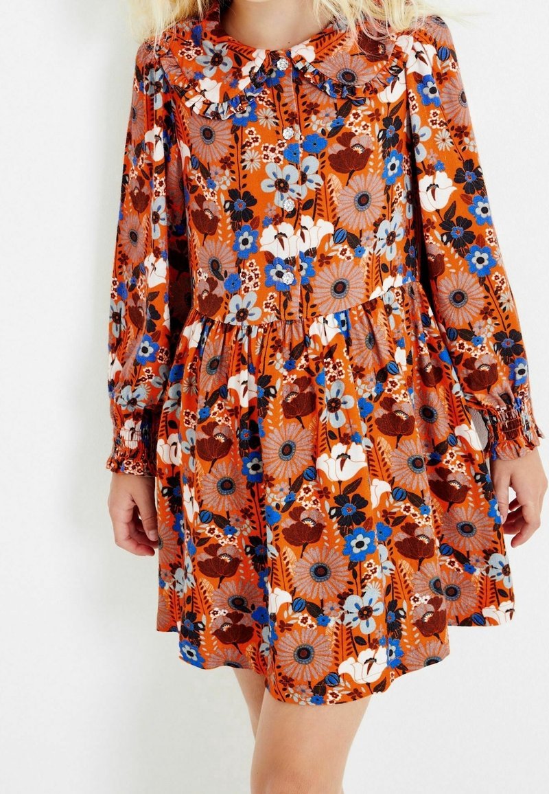 Fille portant une robe orange à manches longues avec des motifs floraux bleus, blancs et bruns et un col à volants, debout devant un fond blanc.
