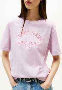 Camiseta de algodón de color morado claro con mangas cortas y escote redondo. Presenta "TOMMY JEANS NEW YORK CITY" impreso en rosa en la parte delantera.