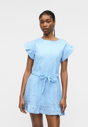 Kvinde iført lyseblå øjenlet kjole med flæseærmer, bundet taljebælte og flæsekant, stående mod en ensfarvet grå baggrund.