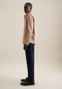Camicia button-up beige chiaro con taschino sul petto, abbinata a pantaloni blu scuro e scarpe nere, in piedi su uno sfondo neutro.