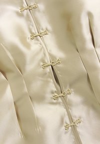 Silk jacket in a light beige color featuring a smooth texture, intricate knotted fastenings, and decorative pleats along the front.