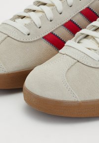 Adidași din suede bej cu dungi zigzag roșii și albastre, șireturi albe și tălpi din cauciuc texturat, având vârful rotunjit și detalii cusute.